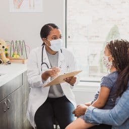 With a protective mask on, a female pediatrician talks to a young patient's mother about the woman's daughter's medical conditions. They are wearing protective masks.