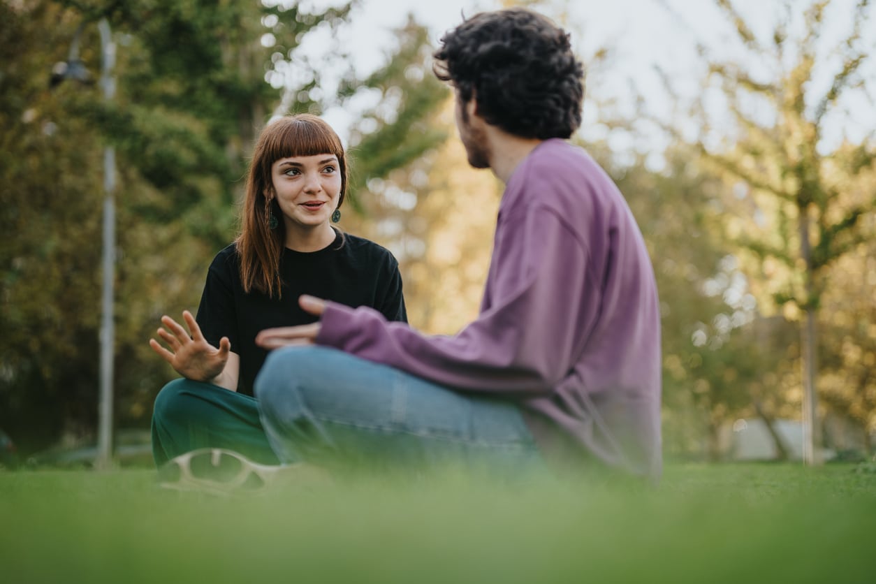 Young friends sitting on grass, enjoying conversation outdoors in bright sunlight.