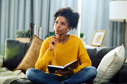 Woman sitting on her couch, reading a book and listening to something through her headphones.